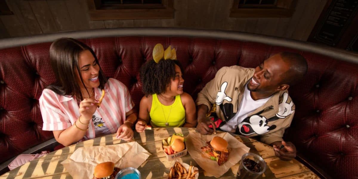 A family of three, including a woman, a young girl, and a man, sit in a red upholstered booth at a restaurant, enjoying burgers and fries. They are all smiling and laughing, creating a joyful atmosphere. The girl is wearing a yellow headband.