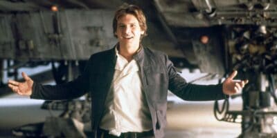 A person stands with a welcoming gesture, arms open wide, smiling. They are wearing a white shirt, dark pants, and a dark jacket. In the background, there is a large, industrial-looking structure with mechanical details.