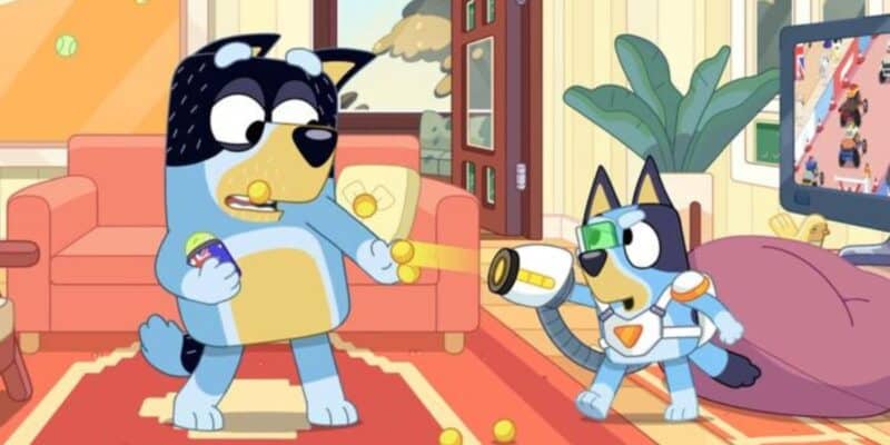 Two animated dogs in a living room, one larger with a black mask and another smaller in a spacesuit, playfully interacting with tennis balls on the floor during a recent episode. Bluey Surprise episode.