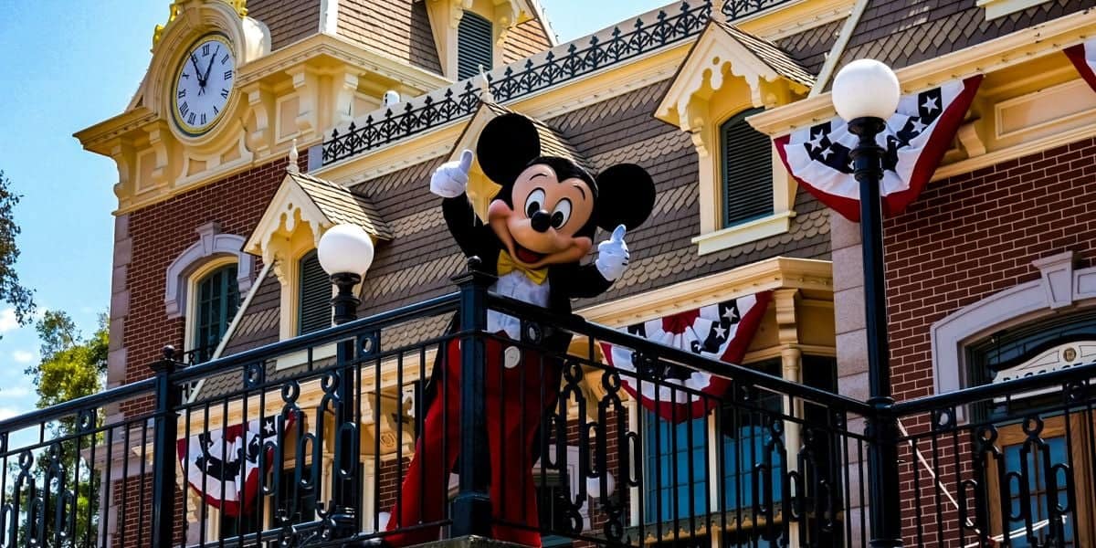 A person in a Mickey Mouse costume waves from a balcony at a theme park. The building behind them features a large clock and decorative elements, including red, white, and blue bunting. As the sky remains clear and blue, visitors are reminded to check for any Disney lost items.