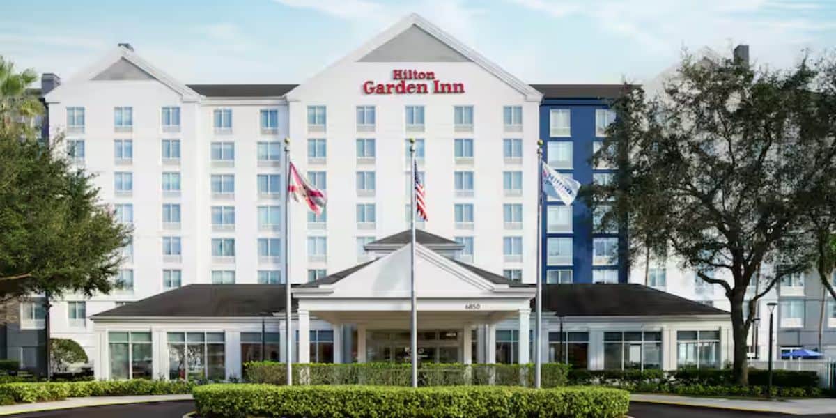 The front view of a Hilton Garden Inn hotel, showcasing a modern white building with a large entrance porch, surrounded by green trees and adorned with multiple flags.