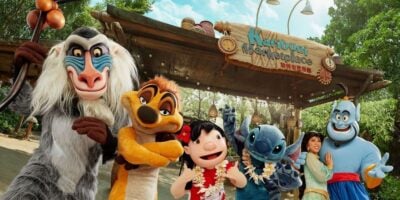 A group of people dressed as Disney characters, including a baboon, a meerkat, Stitch, the Genie, and others, are posing for a photo in front of a sign that reads "Karibuni Marketplace." The background features lush greenery and a wooden structure.