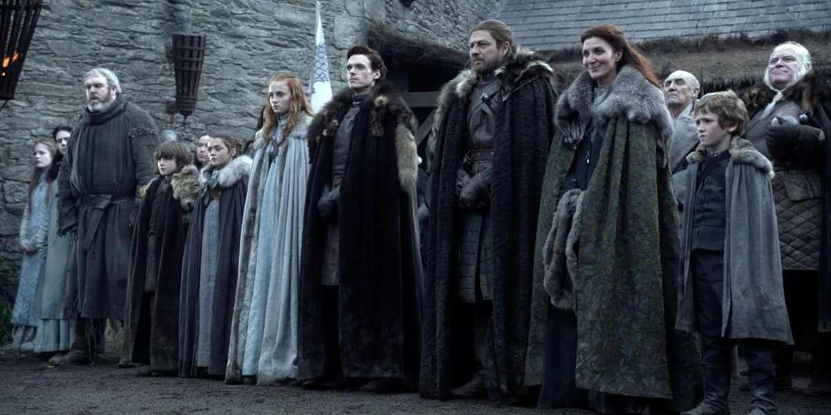 A still from Game of Thrones season one with characters dressed in medieval-style clothing, including furs, stand solemnly in a line against a backdrop of a gray stone castle.