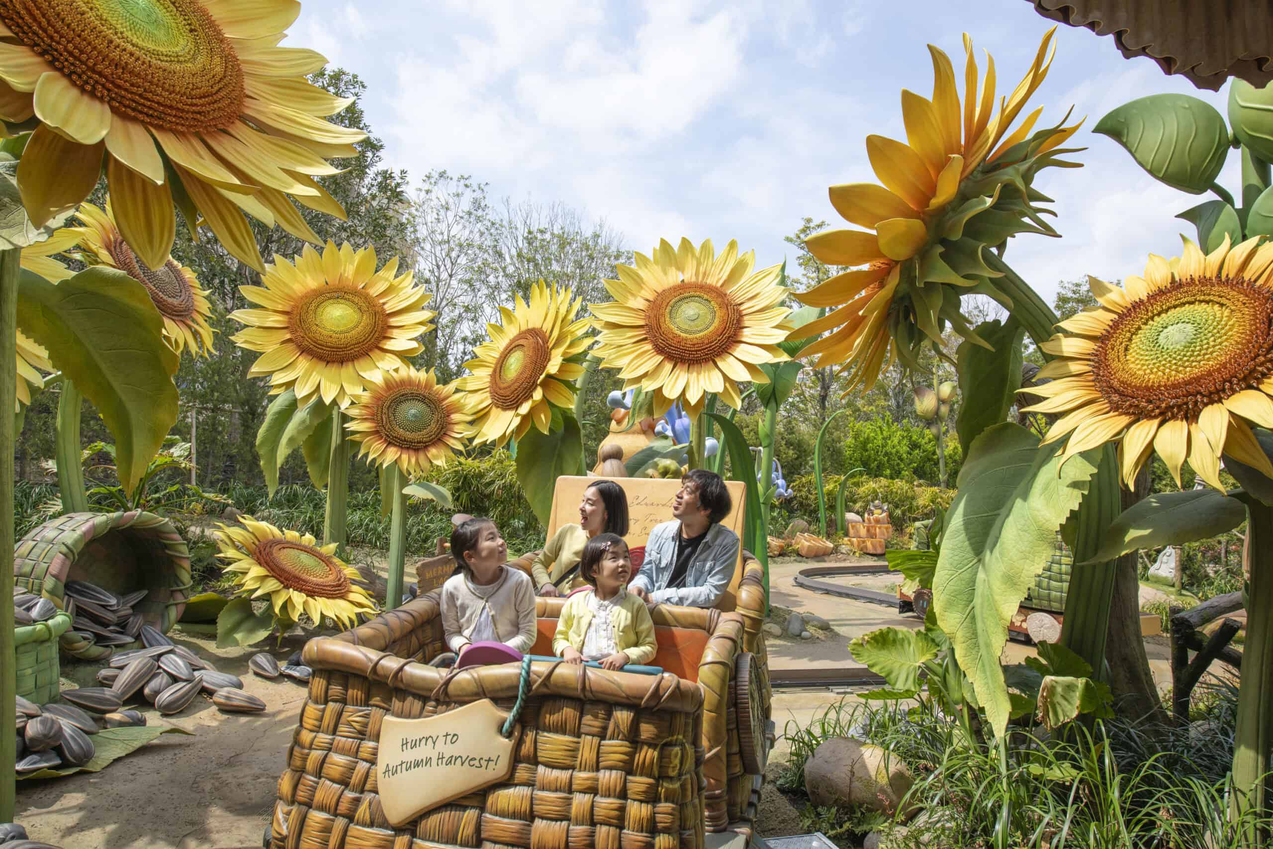 A group of four people, including two children and two adults, sit in a large wicker basket surrounded by oversized sunflowers. The children smile while the adults look fondly at them. A sign on the basket reads "Happy Autumn Harvest!" The scene is bright and cheerful.