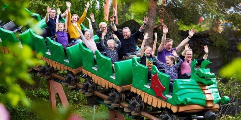 A group of people are enjoying a ride on a green dragon-themed roller coaster at the theme park. They have their hands up and are smiling and laughing. The coaster is surrounded by lush green trees and foliage, suggesting a lively and fun atmosphere at the amusement park.