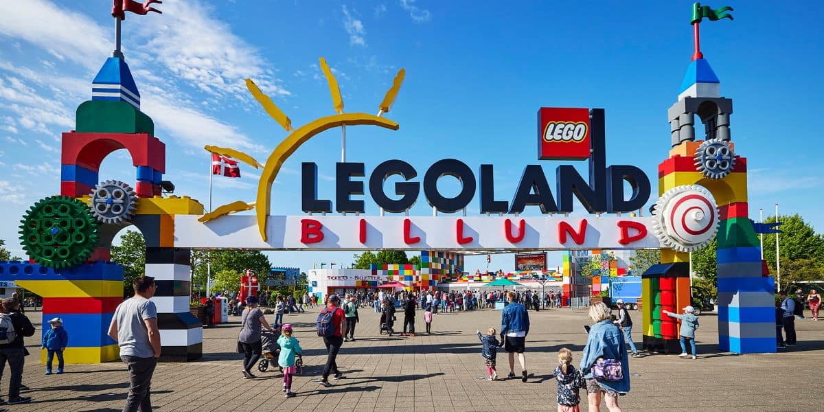 The entrance to Legoland Billund features colorful, LEGO-themed structures with gears and a large sign. A crowd of people, including families and children, are walking towards the theme park entrance. Two Danish flags are seen flying above in the partly cloudy sky.
