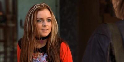 Lindsay Lohan with long, light brown hair wearing a red top and a black choker is talking to someone off-camera, showing a focused expression.