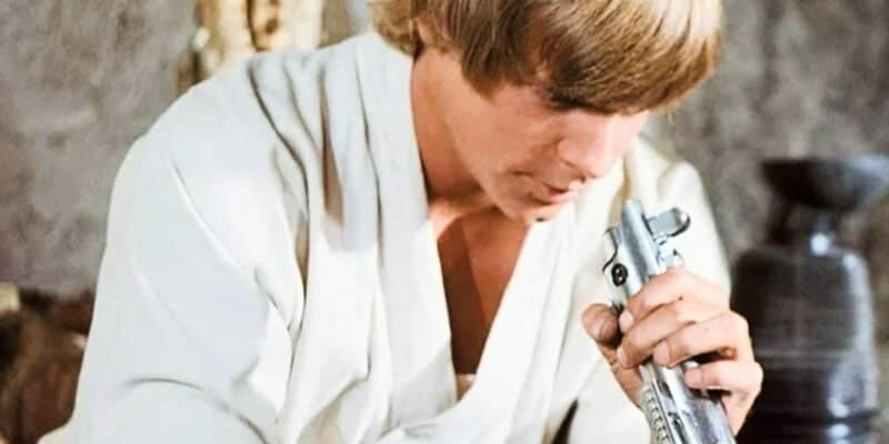 A young man with short blonde hair, dressed in a beige outfit, is looking down at a silver-colored cylindrical object he is holding in his hands. The background is indoors, with blurred elements suggesting a rustic setting.