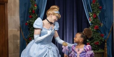 A woman dressed as cinderella interacts warmly with a young girl in a purple princess dress. they are surrounded by elegant curtains and floral decorations at Disney World.