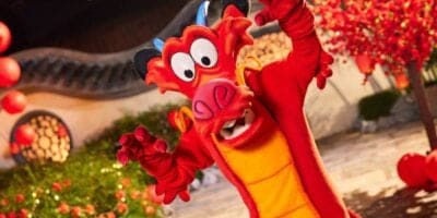 A costumed character of Mushu, a red dragon with a yellow belly, from Disney's "Mulan," is posing energetically. The background features Chinese architectural elements and red lanterns, creating a festive atmosphere.