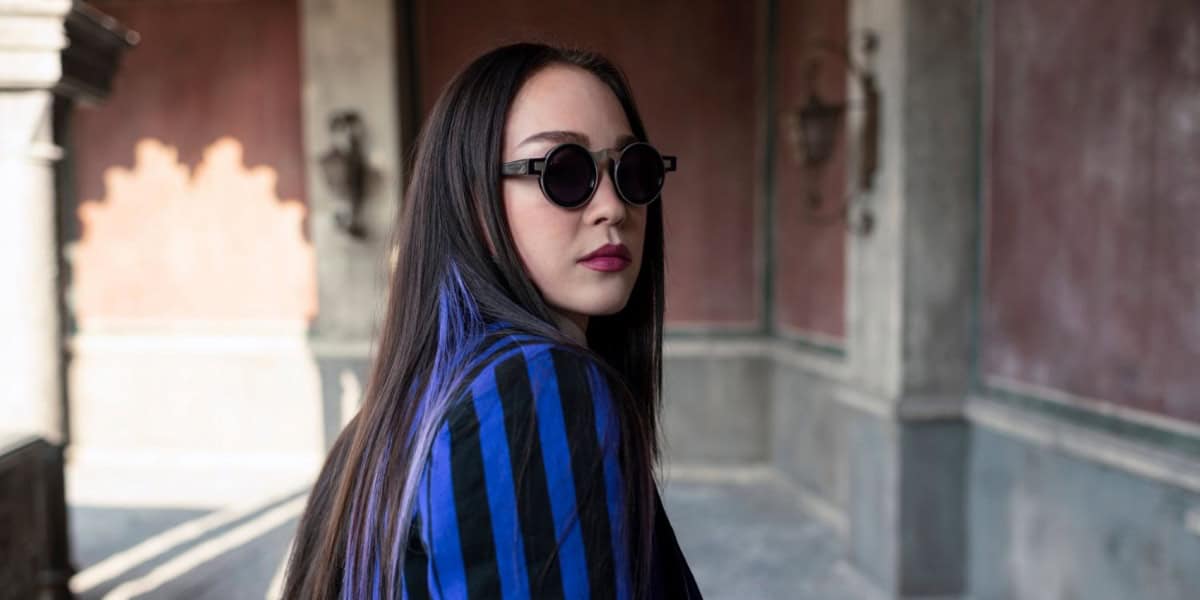 A woman with long dark hair wearing round sunglasses and a blue striped blazer looks back over her shoulder, with a blurred historic building in the background.