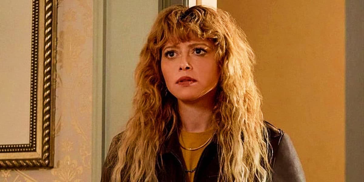Natasha Lyonne with long, wavy blonde hair and bangs stands indoors, looking slightly to the left. They are wearing a brown leather jacket over a yellow top, with a framed picture in the background on the left side of the image.