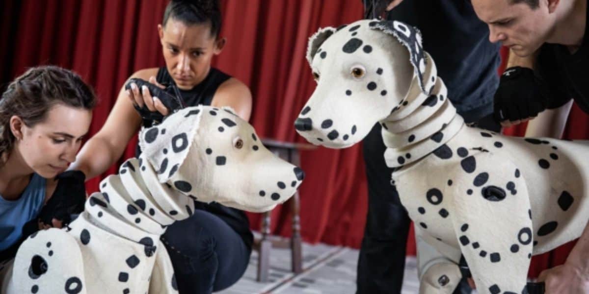 Two performers, each holding black gloves, expertly manipulate life-sized Dalmatian dog puppets covered in white fur with black spots, reminiscent of 101 Dalmatians. The background features a red curtain. Another performer is visible in the distance.