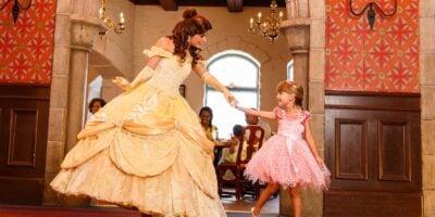 A young girl in a pink dress joyfully meets a woman dressed as a princess in a grand ballroom, both smiling and reaching out hands to each other.