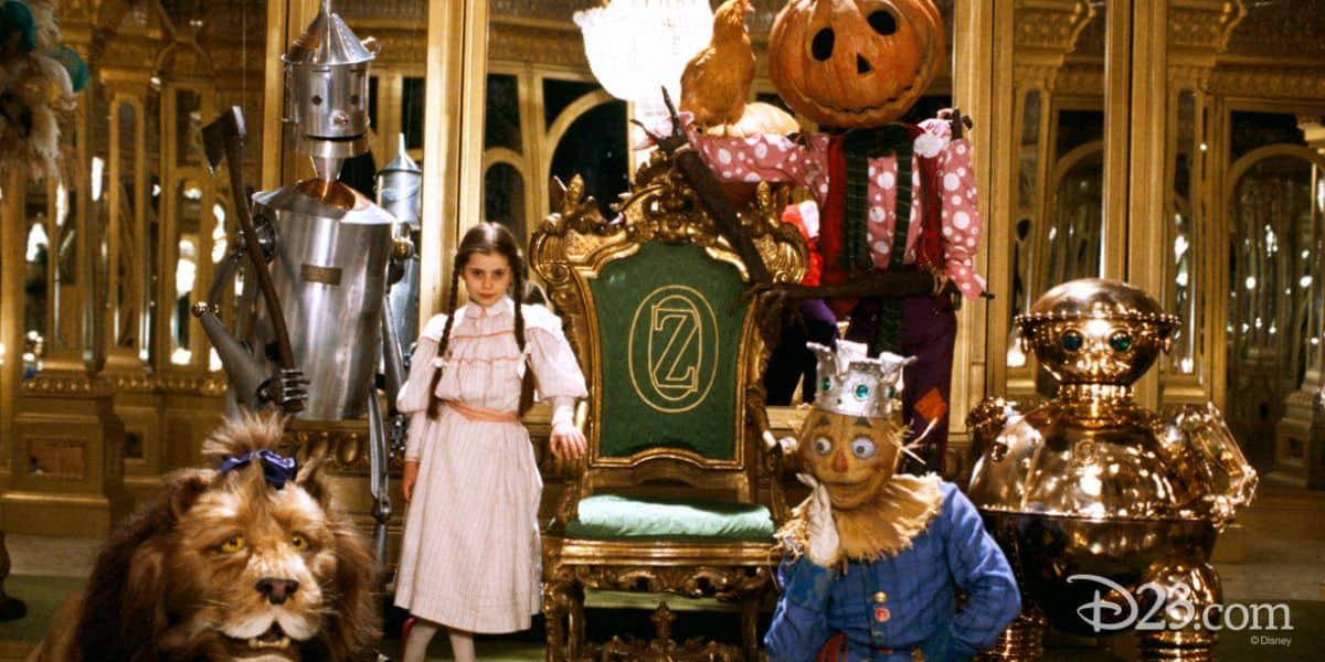 Dorothy and her eclectic group of friends, including a lion, a tin man, a scarecrow, and a robot, stand in a richly decorated palace hall on the set of 'Return to Oz.'