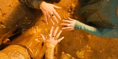 A close-up view of three people extending their hands towards the center, displaying rings. The background is earthy and adorned with fallen leaves. The individuals are dressed in intricate, medieval-style clothing, suggesting a fantasy or historical setting.
