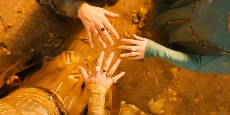 A close-up view of three people extending their hands towards the center, displaying rings. The background is earthy and adorned with fallen leaves. The individuals are dressed in intricate, medieval-style clothing, suggesting a fantasy or historical setting.