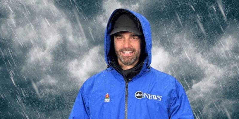 A male news reporter, wearing a blue abc news raincoat and hood, smiling as he reports in a heavy rainstorm.