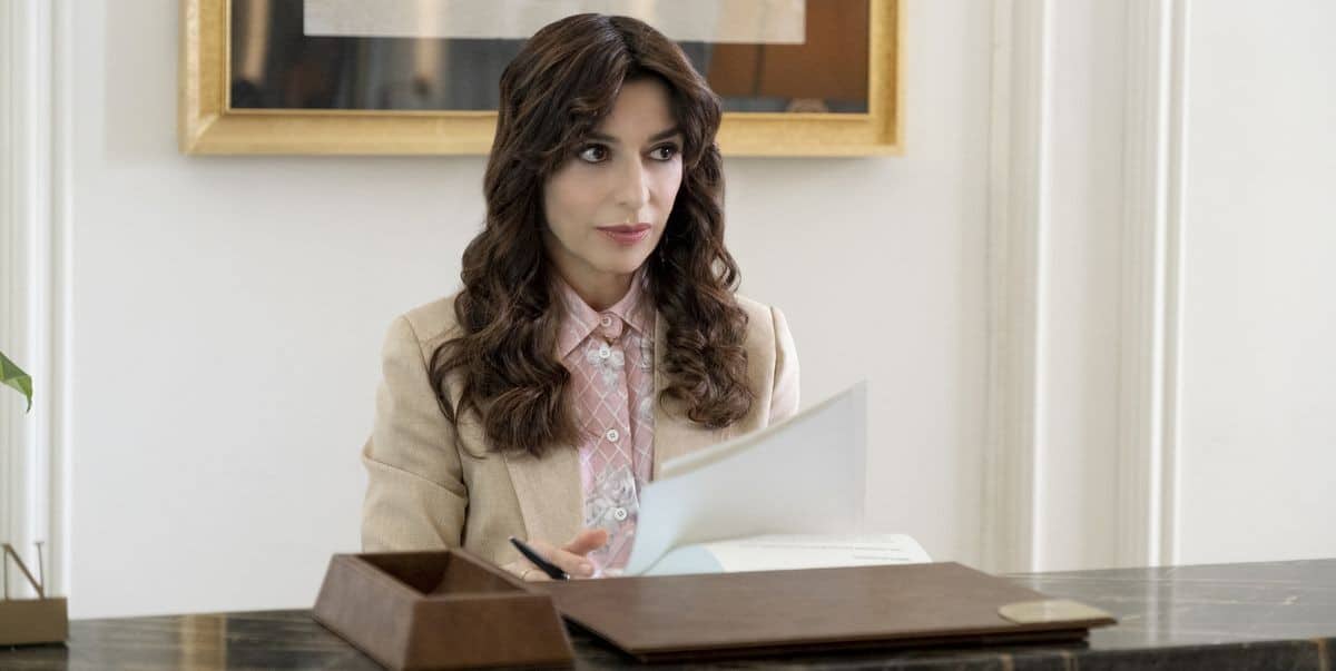 A woman with dark curly hair sitting at a desk in an office, reviewing documents. she is wearing a beige blazer and a pink blouse with a ribbon tie. there is a wood box and a potted plant on the desk.