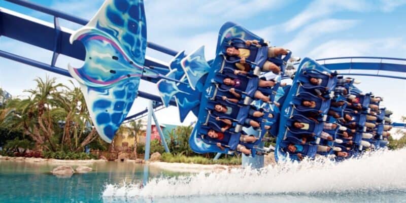 Riders at a Florida theme park on a water roller coaster featuring stingray-themed cars splash down into water beneath a clear blue sky, surrounded by lush tropical vegetation.