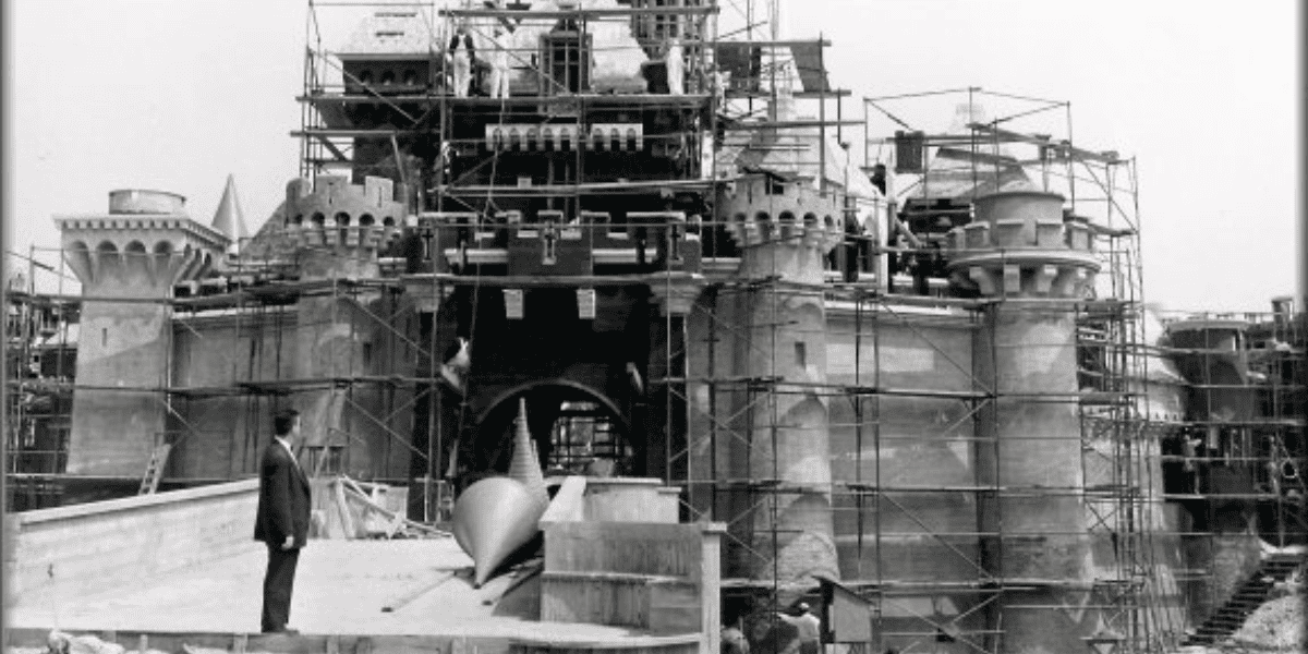 A black and white photograph shows a large castle under construction, surrounded by scaffolding. Workers are on-site, and various construction materials are visible. In the foreground, a man in a suit—reminiscent of Walt Disney—stands observing the progress with keen interest.