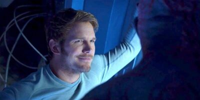 A man with light brown hair and a beard, wearing a light blue long-sleeved shirt, stands in a blue-lit room. He is smiling and leaning against a wall, facing another person whose face is mostly out of frame. Wires can be seen in the background.
