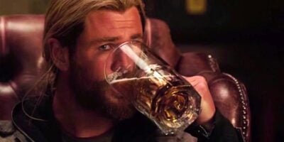 A person with long, light hair and a beard drinks from a large mug of beer while sitting in a leather chair. The atmosphere is cozy and dimly lit, suggesting a bar or pub setting.