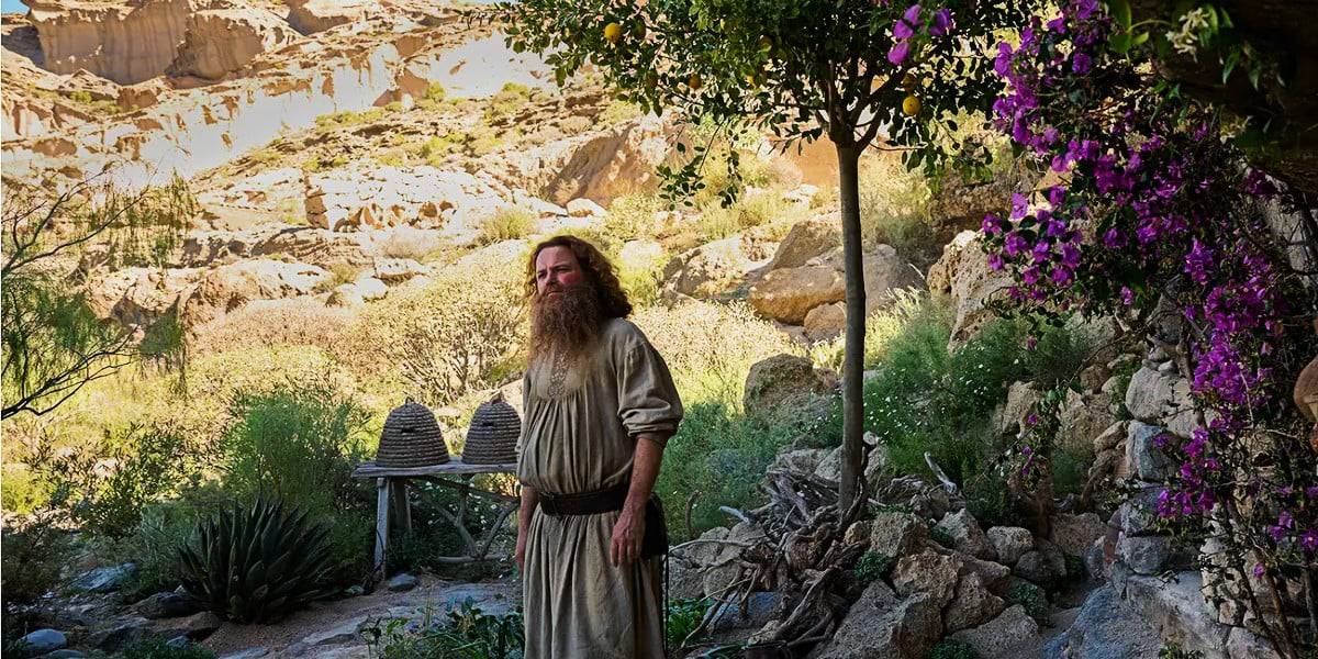 A person with long hair and a beard wearing a robe stands in a rocky, semi-arid outdoor setting. Purple flowers bloom on a tree to the right. There is a stone structure in the background and rugged cliffs under a bright sky. The scene is sunlit and serene.