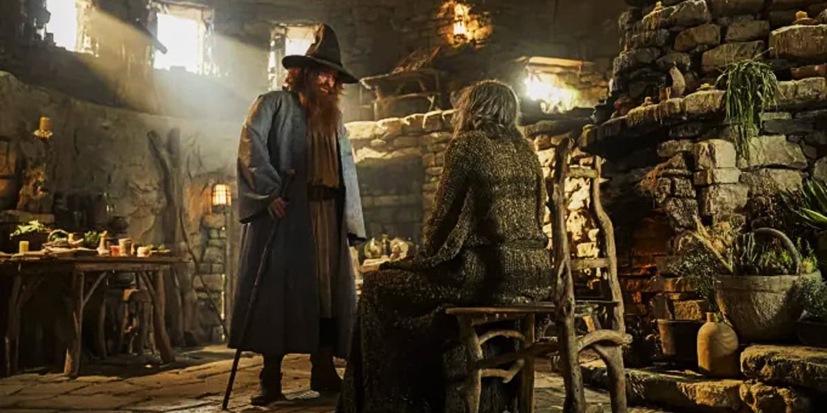 In a dimly lit, rustic stone room, a bearded man in a wizard hat and robe stands next to a seated elderly woman. Various items, plants, and candles adorn the room, contributing to a medieval, magical atmosphere. Sunlight streams in through small windows.
