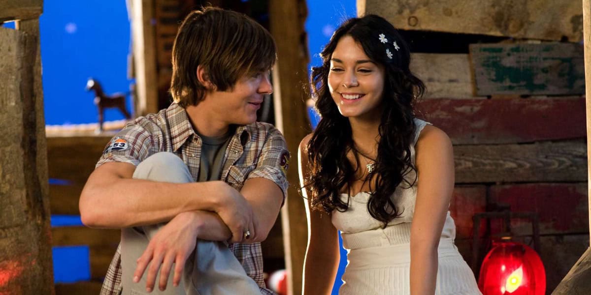 A young man with brown hair wearing a plaid shirt leans against a wooden structure, smiling at a young woman with long dark hair in a white dress, who is also smiling. They are in a rustic setting with wooden planks and a red lantern visible in the background, almost like stepping into a Disney fairytale.