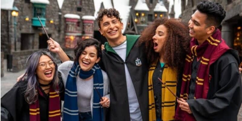 A group of five friends dressed in Harry Potter-themed robes and scarves are posing together, smiling and holding wands. They are standing in front of a backdrop resembling Hogsmeade village from the Harry Potter series. The scene suggests a fun, themed outing at Universal Orlando Resort.