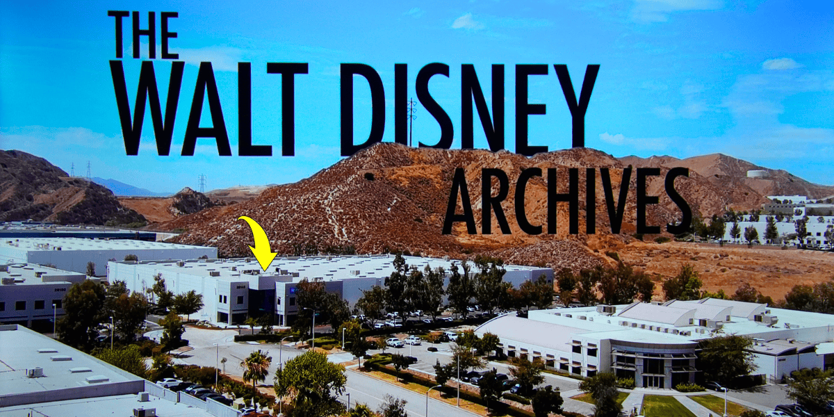 A wide-angle view of a sprawling building complex with "THE WALT DISNEY ARCHIVES" text overlay in large letters. The buildings are set against a backdrop of brown hills under a clear blue sky, reminiscent of an unexpected airport encounter that every Walt Disney fan swears by. A yellow arrow points to one of the buildings in the complex.