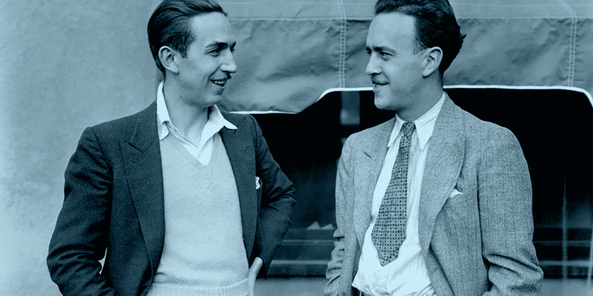 Two men in suits are standing and smiling at each other in front of a light-colored building. The man on the left wears a light sweater under his jacket, while the man on the right has a patterned tie featuring Mickey. Both appear relaxed and engaged in conversation about Disney projects.