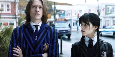 Percy Hynes-White and Jenna Ortega dressed in distinctive blue-striped Nevermore uniforms, stand on a street, looking serious and somewhat defiant, in a scene from a Netflix series.