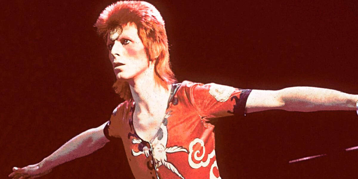 Person with bright red hair and dramatic makeup performing on stage. They are wearing a flamboyant red costume with intricate patterns, striking a pose with arms outstretched. The background is dark, emphasizing the performer's vibrant appearance.