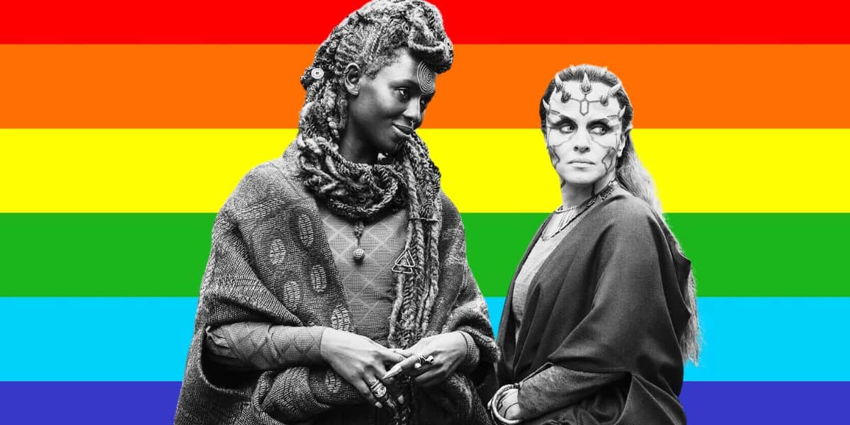 Two people dressed in elaborate, tribal-inspired costumes, reminiscent of characters from "The Acolyte" series, stand engaging in conversation. The black-and-white image has a colorful rainbow flag as the background, evoking a sense of unity akin to an iconic "Star Wars" Episode 3 scene.