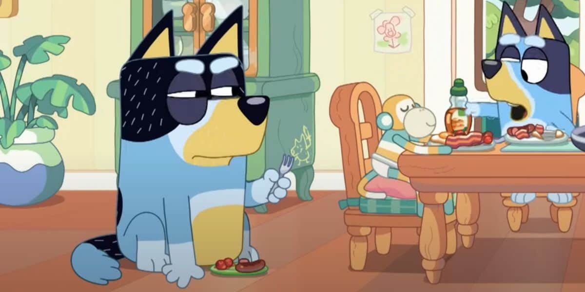 A blue and black cartoon dog, reminiscent of Bluey, sits on the floor holding a fork with food, looking unimpressed. In the background, another blue and black cartoon dog and a small character sit at a dining table with food. The room, designed by talented animators, has green plants and a painting on the wall.