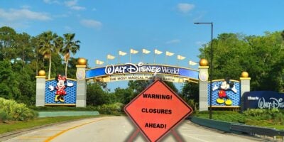 A road leading to the entrance of Walt Disney World, featuring colorful signs with Mickey and Minnie Mouse on either side. In the foreground, a bright orange warning sign reads "Warning! Closures Ahead!" against a backdrop of Disney-themed trees and a clear blue sky.