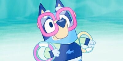 Animated character Bluey, a blue heeler puppy, stands in a pool with a joyful expression. She wears large pink goggles, floaties on her arms, and a dark blue swimsuit with a fish design. The background perfectly captures the essence of summer fun with its soft blue-green color scheme.