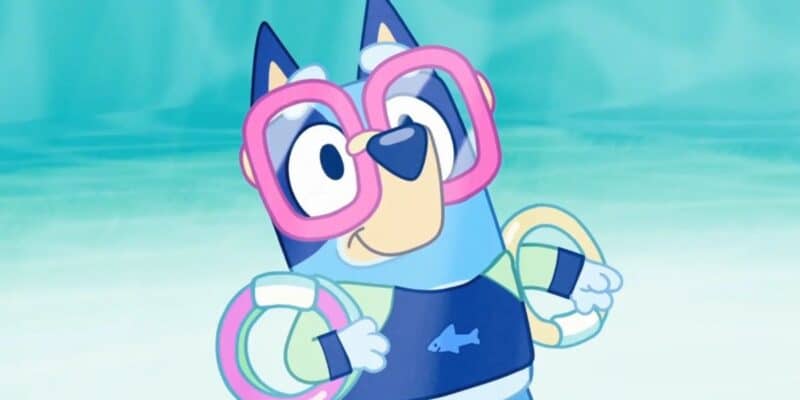 Animated character Bluey, a blue heeler puppy, stands in a pool with a joyful expression. She wears large pink goggles, floaties on her arms, and a dark blue swimsuit with a fish design. The background perfectly captures the essence of summer fun with its soft blue-green color scheme.