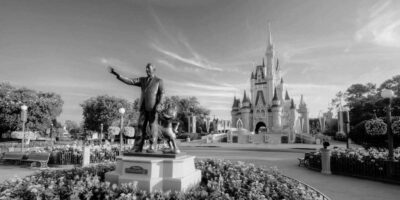 Black and white photo of a classic Disney scene featuring a statue of a man holding hands with a mouse character in the foreground. The statue stands in a landscaped area with flowers, evoking memories of a magical Disney World trip, while a fairytale-like castle is prominent in the background.