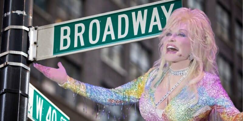 A person with long blonde hair, dressed in a colorful, sparkly outfit reminiscent of Dolly Parton, sings with their arms outstretched in front of a Broadway street sign at the corner of W 44th St. The background shows a building facade with multiple windows.