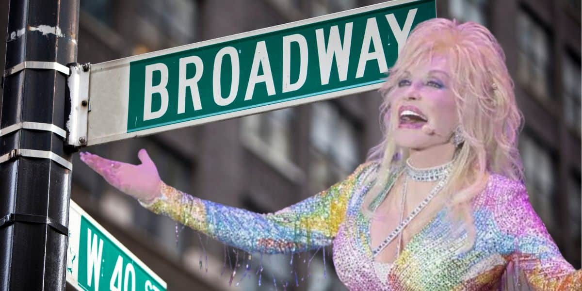 A person with long blonde hair, dressed in a colorful, sparkly outfit reminiscent of Dolly Parton, sings with their arms outstretched in front of a Broadway street sign at the corner of W 44th St. The background shows a building facade with multiple windows.
