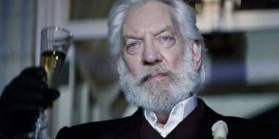 An elderly man with white hair and a beard is holding a glass of champagne. He is dressed formally in a dark suit and white shirt, with a white flower pinned to his lapel. The background is slightly blurred, suggesting an auto draft effect, and he has a serious expression on his face.