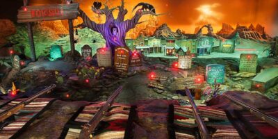 An amusement park shooting gallery with a spooky Halloween theme debuts as a controversial attraction on Opening-Day. The display features a purple tree, gravestones with humorous inscriptions, faux shotguns in the foreground, and an eerie, glowing orange backdrop.