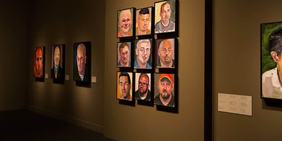 A dimly lit gallery features multiple portraits hung on green walls. One wall showcases a 4x4 grid of colorful, expressive portraits, each with unique subjects. Additional framed paintings hang on the adjacent wall to the left, with a small plaque visible between them.