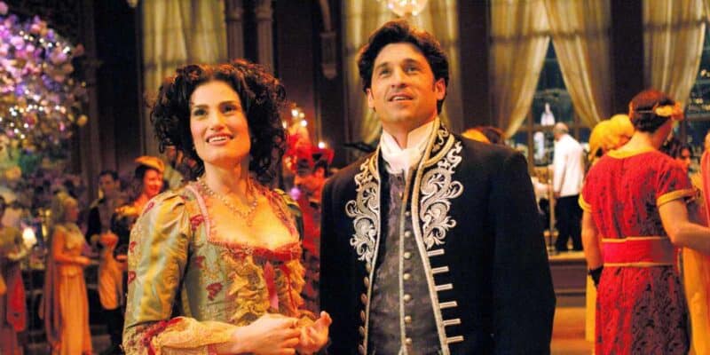 Two people, a woman and a man, dressed in elegant period costumes at an enchanted, lavishly decorated ballroom. The woman wears a gold and red gown with intricate details, while the man sports a black jacket with silver embroidery. They both appear to be admiring the surroundings.