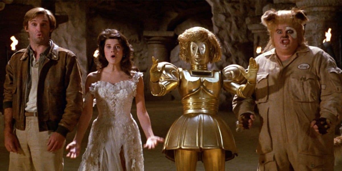 A group of four characters stands in a cave-like setting. From left to right: a man in casual clothes, a woman in a white dress, a gold humanoid robot reminiscent of Spaceballs’ C-3PO spoof, and a man in a bear-like costume. All characters have surprised expressions on their faces.