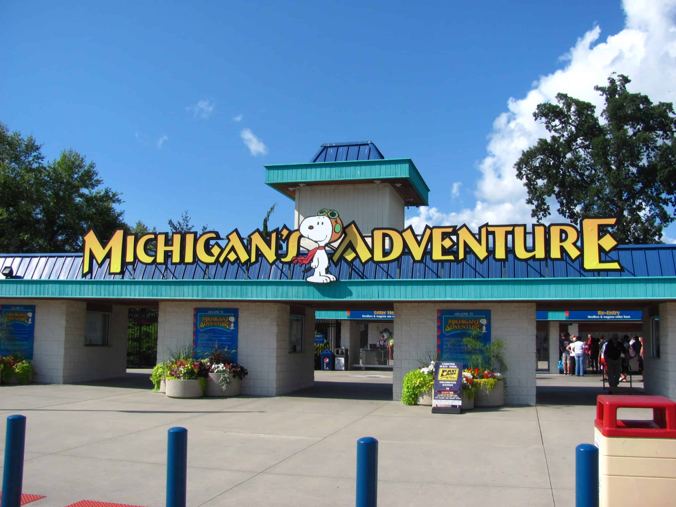 Entrance of Michigan's Adventure amusement park with the park's name displayed prominently, featuring a character from the Peanuts comic strip. The entrance has ticket booths and decorative plants, set against a bright blue sky with scattered clouds.