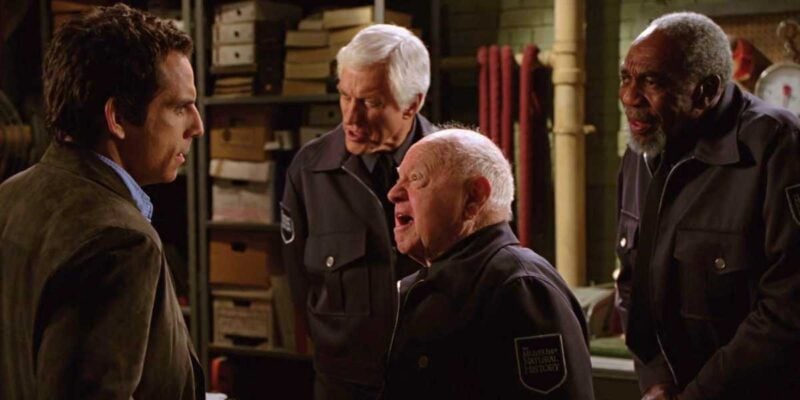A man in a brown jacket converses with three older men wearing security guard uniforms in what appears to be a storage or maintenance room. Shelves with various items and equipment can be seen in the background. The men are engaged in a lively discussion.